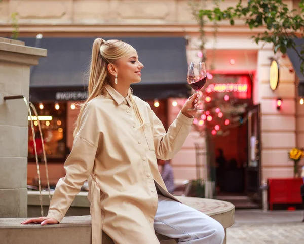 古いヨーロッパの都市の味の夜の通りにベージュのコートで魅力的な若い女性と赤ワインを楽しむ — ストック写真