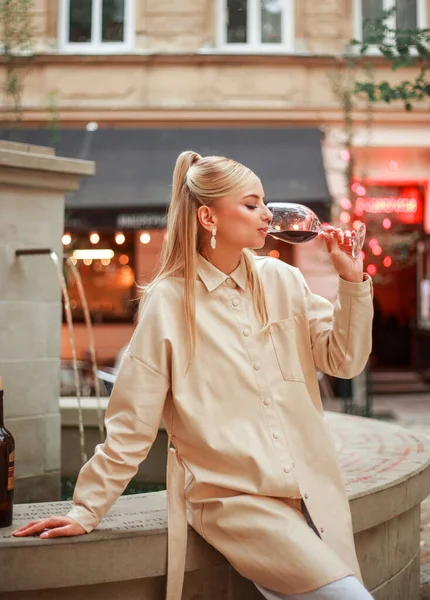 Eski bir Avrupa şehrinin akşam caddesinde bej ceketli, alımlı genç bir kadın kırmızı şarabı tadıyor ve seviyor.