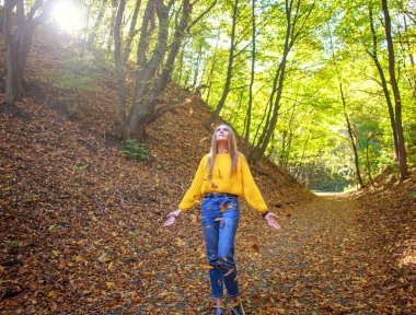 Sırt çantalı genç bir kadın şehir parkında yürüyor ve güneşli bir sonbahar gününde doğanın tadını çıkarıyor.