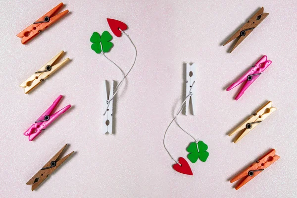 Colorful set of different wooden and plastic clothes pegs on a light pink glitter background. A red and a green lucky clover leaf are attached to each of the white wooden clothes pegs. Top view.