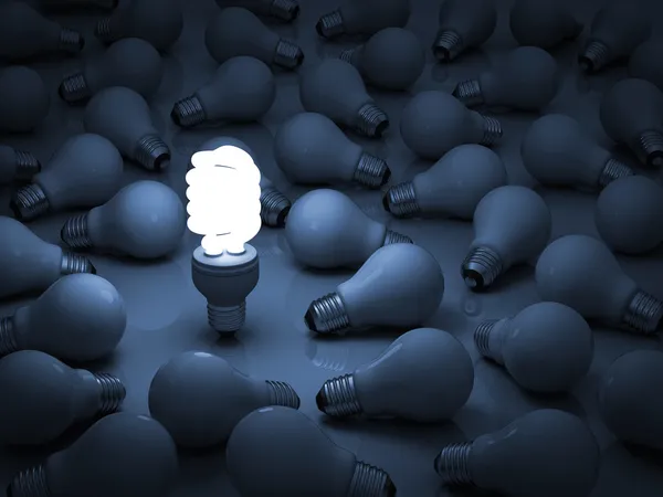 One glowing eco compact fluorescent light bulb standing out from the unlit incandescent light bulbs — Stock Photo, Image