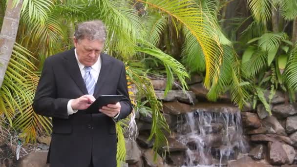 Mogna affärsman står med sin tablett genom ett vattenfall med palmer. — Stockvideo