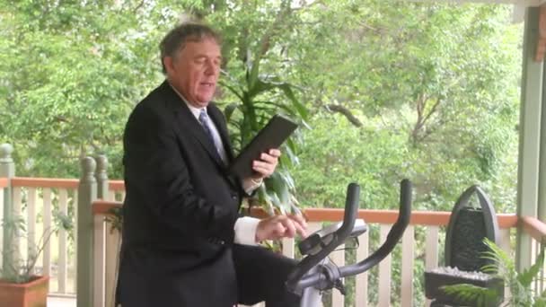 Mature businessman working out on an exercise bike while working with his tablet. — Stock Video