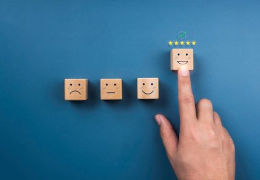 Müşteri hizmetleri değerlendirmesi, reytingler, geri bildirim, müşteri deneyimi ve memnuniyet anketleri. Beş yıldızlı işaretler ve mavi arkaplandaki tahta küp bloğundaki mutlu yüzü gösteren el işaretleri.