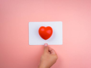 Kırmızı kalp topu beyaz kare kağıtta, yuvarlak köşeler pembe pastel arka planda elle tutuluyor. Sevgi, ilgi, paylaşım, refah ve bağış kavramları, asgari stil, minimal stil.