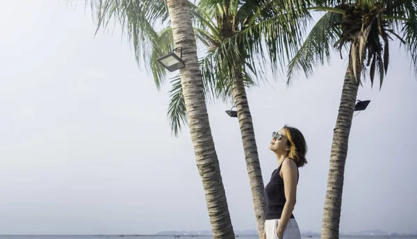 亚洲女人看上去很酷 留着短发 戴着太阳镜 穿着黑色的背心上衣和白色的裤子 双手插在海滩上椰子树下的口袋里 还有大海的背景 — 图库照片