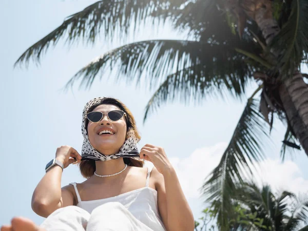 身穿休闲白衬衫 戴着太阳镜和发巾的美丽而快乐的亚洲女人画像 夏天在椰子树下的沙滩上 蓝天背景下 带着微笑享受着 — 图库照片