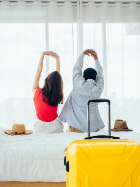 Birkaç turist, mutlu tatiller. Yaz tatili. Genç Asyalı kadın ve erkeğin, otel odasındaki pencerenin perdesinin yanındaki bavulla yatakta gerinirken, dikey bir şekilde..