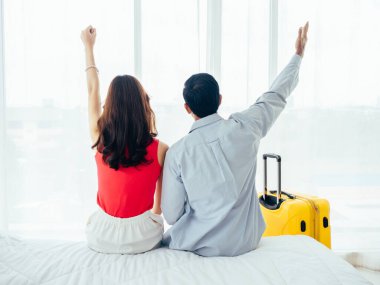 Birkaç turist, mutlu tatiller. Yaz tatili. Genç Asyalı erkek ve kadının arka plandaki portresi beyaz yatakta mutlu bir şekilde el kaldırırken otel odasının penceresinin yanındaki perdenin yanında bavulla..