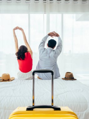 Birkaç turist, mutlu tatiller. Yaz tatili. Genç Asyalı kadın ve erkeğin, otel odasındaki pencerenin perdesinin yanındaki bavulla yatakta gerinirken, dikey bir şekilde..