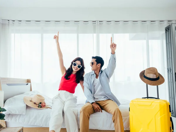 Ready to travel, happy holiday, check-in. Summer vacation concept. Asian couple pointing finger up with joy, man and woman wear sunglasses smile with happy on white bed with yellow suitcases on trip.