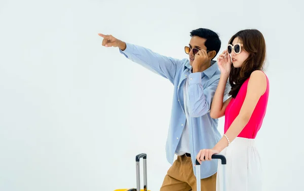 Ready to travel, happy holiday. Summer vacation. Asian couple in sunglasses with pointing and looking forward with exciting, young woman and man with suitcase on trip, isolated on white background.