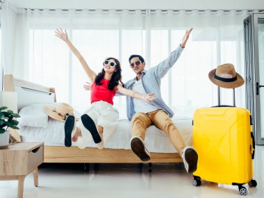 Yolculuğa hazır, iyi tatiller, giriş. Yaz tatili kavramı. Asyalı çift sevinçle el kaldırıyor, erkek ve kadın güneş gözlüğü takıyor mutlu bir şekilde beyaz yatakta, sarı bavullarla geziyorlar..