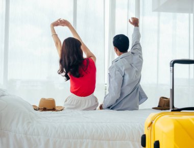 Birkaç turist, mutlu tatiller. Yaz tatili. Genç Asyalı kadın ve erkeğin arka plandaki portresi otel odasının penceresindeki perdenin yanındaki bavulla beyaz yatağa uzanıyor..