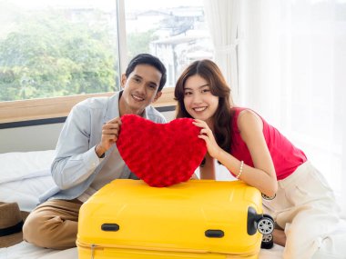 Çekici Asyalı sevgili balayı gezisine hazır, yaz tatili tatili. Mutlu yakışıklı adam ve güzel gülümseyen kadın beyaz odada kırmızı bir kalp yastığı ve sarı bir bavul tutuyor..