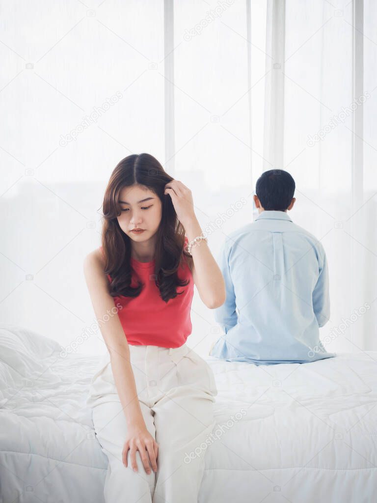 The anxiety of Asian couple lovers on the bed. Sad young woman and man sitting on the bed with relationship difficulties feeling sad and thinking in the bedroom at home near the glass window.