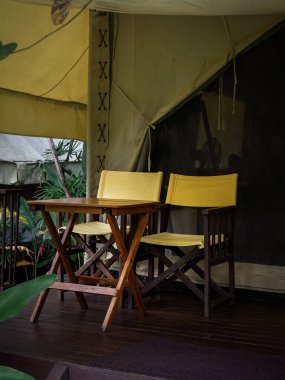 İki boş kamp ahşap koltuğu, sarı kumaş koltuklar, sırt sehpası ve büyük çadırın önündeki terasta ahşap masa. Beyaz glamping çadırında katlanır sandalyeler, dikey stil.