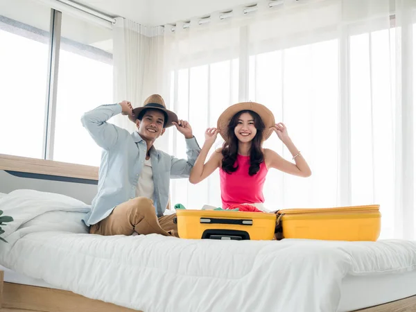 Couple travelers ready to trip. Young Asian woman and man in denim shirt wearing beach hats while packing with yellow suitcases in bedroom, looking at camera. Colorful summer vacation. Happy holiday.