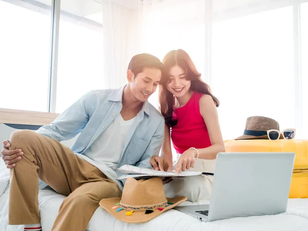 Ready to trip and vacation plan concept. Young pretty Asian woman and man in denim shirt pointing fingers on the map near laptop on the bed with suitcase. Travel planning on summer holiday.