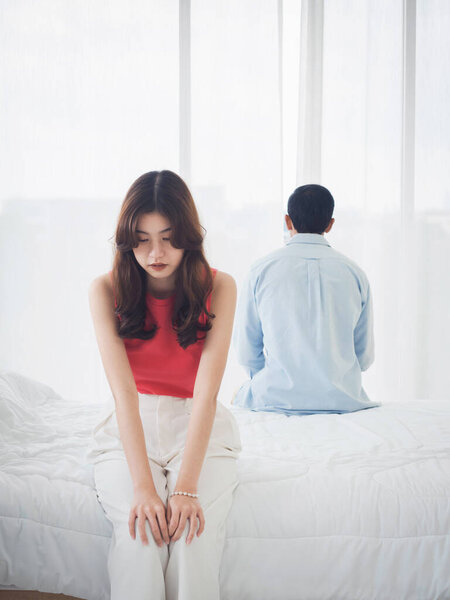 The anxiety of Asian couple lovers on the bed. Sad young woman and man sitting on the bed with relationship difficulties feeling sad and thinking in the bedroom at home near the glass window.