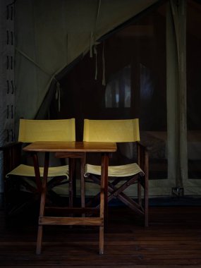 İki boş kamp ahşap koltuğu, sarı kumaş koltuklar, sırt sehpası ve büyük çadırın önündeki terasta ahşap masa. Beyaz glamping çadırında katlanır sandalyeler, dikey stil.