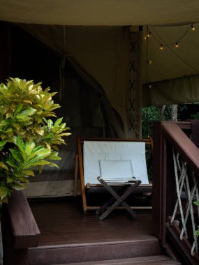 Büyük çadırın önündeki terasta beyaz kumaş koltuklu ve ayaklı boş bir kamp koltuğu. Beyaz glamping çadırı üzerinde katlanır güneş yatağı, dikey stil.