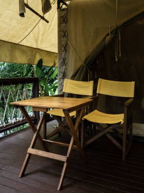 İki boş kamp ahşap koltuğu, sarı kumaş koltuklar, sırt sehpası ve büyük çadırın önündeki terasta ahşap masa. Beyaz glamping çadırında katlanır sandalyeler, dikey stil.