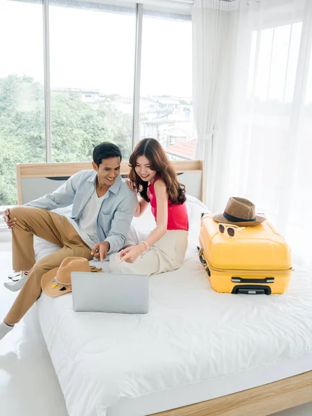 Ready to trip and vacation plan concept. Young pretty Asian woman and man in denim shirt looking at laptop on the bed with suitcase and beach hats, vertical style. Travel planning on summer holiday.