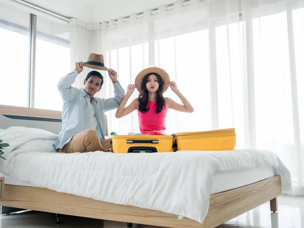 Couple travelers preparing to trip. Young Asian woman and man in denim shirt wearing beach hats while packing with yellow suitcases on bed in the bedroom. Colorful summer vacation. Happy holiday.