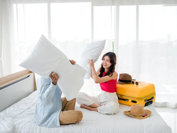 Couple pillow fight. Asian beautiful young woman and man in denim shirt sitting and fighting together with pillows near the yellow suitcase on the bed in the bright hotel room. Happy holiday.