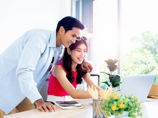 Happy Asian couple, young woman and man using laptop computer on desk together for flight booking, hotel room and trip information in office, ready to travel, happy holiday, summer vacation concept.