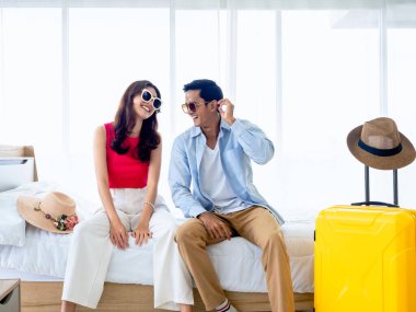 Yazın mutlu bir Asyalı çift, güneş gözlüğü takan genç bir adam ve kadın, gülümseyen bir güneş gözlüğü takıyor, valiz ve plaj şapkasının yanındaki parlak yatak odasında oturuyor, tatil seyahati, yaz tatili konsepti.