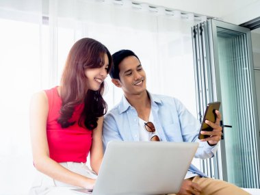 Mutlu Asyalı çift, genç adam ve kadın gündelik hayatlarında akıllı cep telefonu ekranlarına bakarken aynı anda dizüstü bilgisayarla çalışıyorlar ve arkadaşlarıyla aydınlık odada video görüşmesi yapıyorlar..