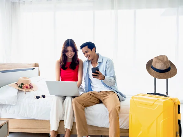 stock image Happy Asian couple, young man and woman looking and using laptop computer and smartphone together for flight booking and trip information in bedroom near baggage, travel on holiday, summer vacation.
