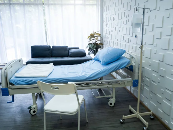 Empty electric adjustable patient bed in a hospital room with blue pillow and bedsheet, saline solution bags, chair, sofa near the window in recovery room, Health care, medical and insurance concept.