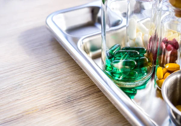 Close up vitamins and supplements in the small clear glass bottles in the steel medical tray on wooden table with copy space. Green, yellow, red, white. Healthcare, and medical concepts.