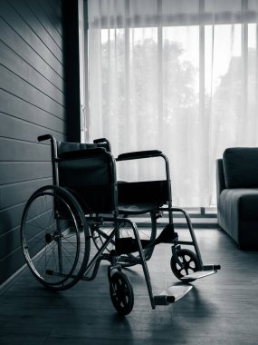 Empty black wheelchair near the blue wall and grey with white curtain at glass window in the living room vertical style, waiting for patient services. Lonely wheelchair with nobody in hospital room.