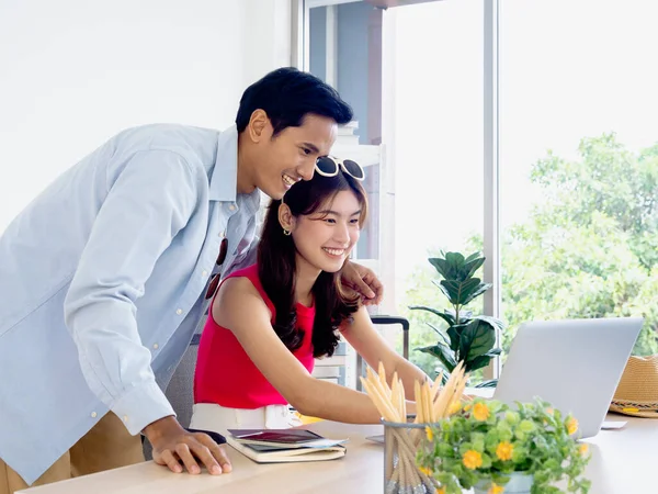 Asian couple, young woman and man looking at laptop computer screen on desk together for travel booking, flight, hotel room and trip information in office, happy summer holiday concept.