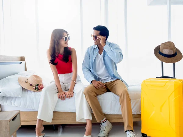 Happy Asian couple in summer, young man and woman wearing sunglasses with smile, sitting on the bed in the bright bedroom near yellow luggage and beach hat, holiday travel, summer vacation concept.