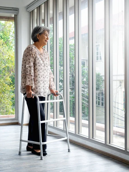 Asian senior woman, white hair standing with walking frame, full length, looking out the glass window, vertical style. Elderly lady patient using walking frame. Strong health, medical care concepts.