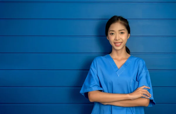 Portrait of friendly, cheerful, smiling confident Asian woman doctor or nurse in blue suit stand with arms crossed on wooden wall background, healthcare professional in blue scrubs with copy space.