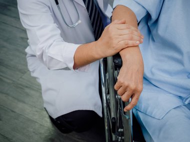Close Up doctor's hands holding elderly patient 's hand in a wheelchair. Male doctor in white suit visit and encourage senior woman patient dressed in light blue in hospital. Elderly care concept.