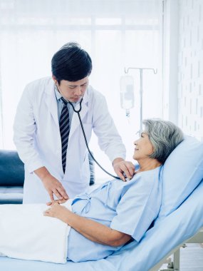 Asian elderly female patient dressed in light blue, smiling in bed while male doctor in white suit uses stethoscope to listen to heartbeat for check-up in recovery room in hospital, vertical style.
