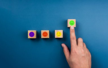 Feedback, client survey, The green one with happy smile face emoticon on wood cube block chosen by hand from other faces on blue background, customer experience satisfaction rating with emotion face.