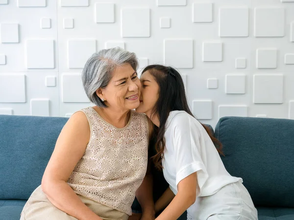 Happy Asian senior, mother white hair embracing her beautiful daughter while she kiss her cheek with love, care, comfort and smile while sit on sofa on white background in living room at home.