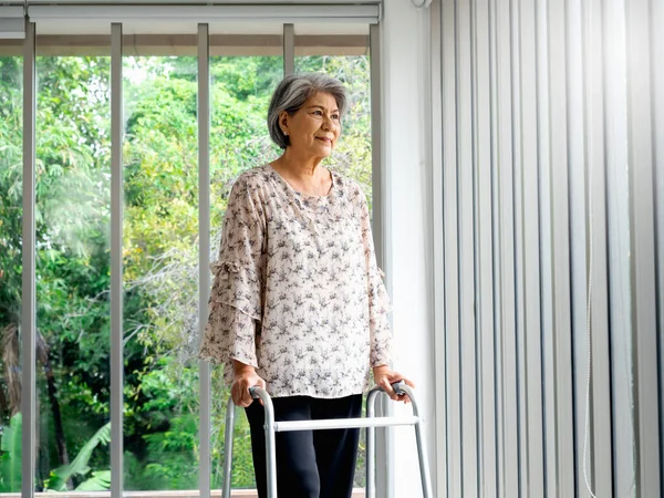 Asian senior woman, white hair standing with walking frame and looking out the glass window, indoors. Elderly lady patient using walking frame. Strong health, medical care and life insurance concepts.