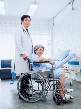 Asian elderly female patient dressed in light blue, smiling happily sits in wheelchair with kindly male doctor in white suit standing beside her  near bed in recovery room in hospital, vertical style.