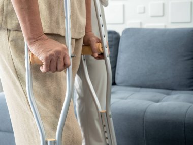 Closeup elderly woman's hands holding on crutches standing in living room at home, trying to walk. Helping and care senior patient to walk with crutches walker concept.