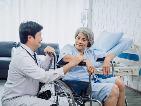 Asian senior woman patient dressed in blue, smiling happily sits in wheelchair with kindly man doctor in white suit holding her hand beside in recovery room in hospital. Healthcare support concept.