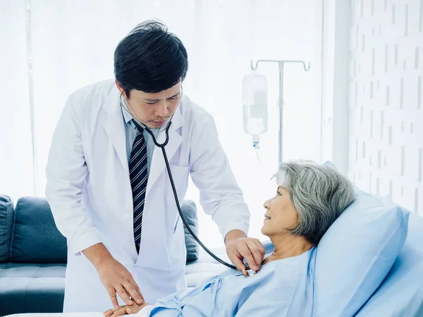 Asian elderly female patient dressed in light blue, smiling happily in bed while the kindly male doctor in white suit uses stethoscope to listen to heartbeat for check-up in recovery room in hospital.